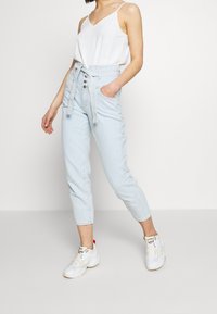 Ljusblå högmidjade jeans med bälte i midjan, knappstängning framtill och avsmalnande ben, kombinerade med en vit linne med smala axelband och vita sneakers.