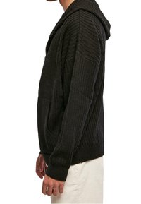 Svart huva med ribbstickad tröja med texturerad yta, med en känguruficka och åtsittande mudd, sedd från sidan.