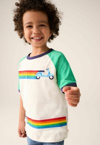 Baltā T-kreklā ar īsām piedurknēm, zaļiem raglan piedurkņu apakšiem. Priekšpusē un apakšējā apmalē ir zila skūtera dizains un varavīksnes svītras.