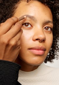 Crayon à sourcils marron appliqué sur le sourcil d'une femme. Elle a des cheveux noirs bouclés et une boucle de septum, avec une peau lisse et un maquillage neutre.