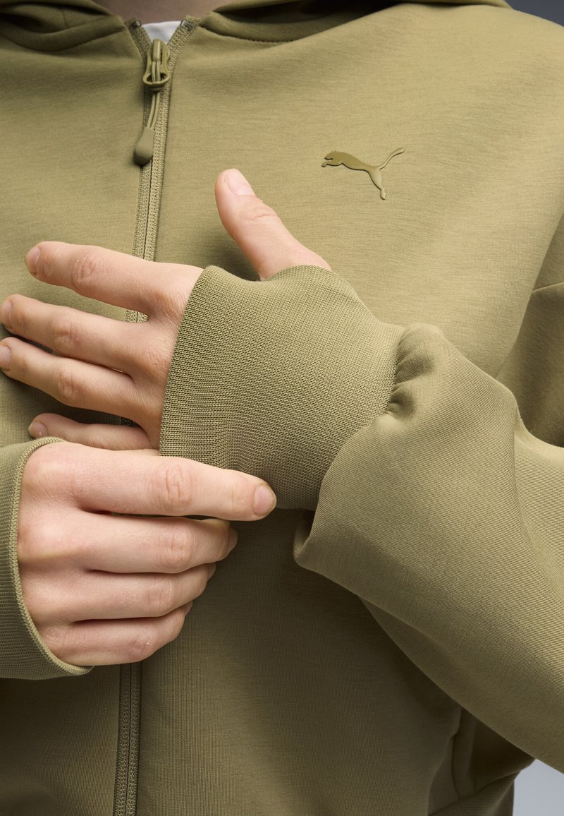Personne portant un sweat à capuche Puma zippé vert olive avec des passe-pouces, ajustant la manche avec la main gauche près de la poitrine.
