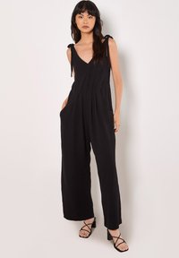 Black, sleeveless jumpsuit with a v-neck, tied shoulder straps, side pockets, and a relaxed fit, featuring a crinkled texture and wide legs.