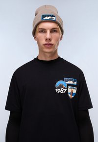 Sort t-shirt med grafiske lapper, der viser bjerglandskaber og året "1987." Beige strikket beanie med et logo-patch i blå og hvid.