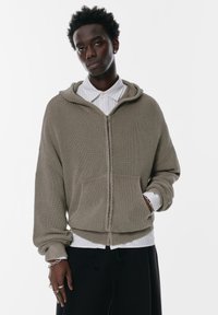 Khaki zipová mikina s žebrovanou texturou, oversized designem a předními kapsami. V kombinaci s bílou košilí pod ní.
