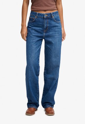 Persoon die blauwe jeans met een hoge taille en rechte pijpen draagt, gecombineerd met bruine schoenen met vierkante neus en een bruin cropped top tegen een witte achtergrond.