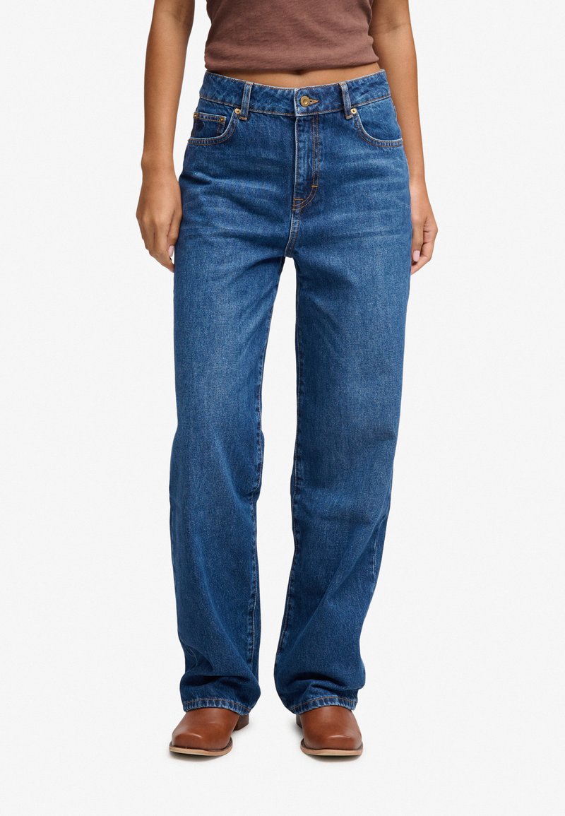 Person, der bærer højtaljede blå jeans med lige ben kombineret med brune sko med firkantet tå og en brun cropped top mod en hvid baggrund.