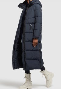 Marinblå vadderad lång kappa med huva, med dragkedjor och en figursydd silhuett. Matchas med vita snörade kängor.