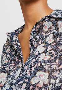 Luftig, genomskinlig blus med blommönster i rosa, vitt och blått mot en mörk bakgrund. Knappslå framtill och detaljer i kragen synliga.