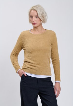 Donna bionda che indossa un maglione peloso color sabbia sopra una camicia bianca, con pantaloni neri, in piedi con una mano in tasca davanti a uno sfondo semplice.