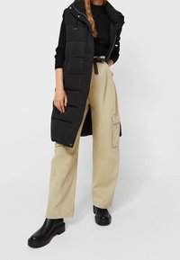 Black puffy sleeveless vest over a black ribbed sweater, paired with light beige cargo pants and black chunky ankle boots.