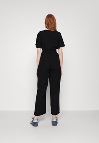 Mono negro con mangas cortas, ajuste holgado, cintura ceñida y piernas rectas. Se lleva con zapatos azules y grises. Diseño simple y sin adornos.