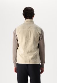 Beige fleece vest with high collar, snug fit, and contrasting brown long sleeves. Smooth texture, simple design, and no visible fastenings.