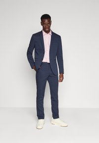 Veste de costume marine et pantalon, chemise rose, baskets blanches ; présente une coupe ajustée, une texture lisse et des détails subtils sur la veste.