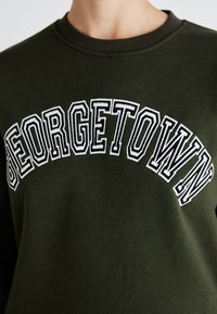 Temno zelena kapuca z belim vezenim napisom "GEORGETOWN", okrogel izrez in mehak bombažen material.