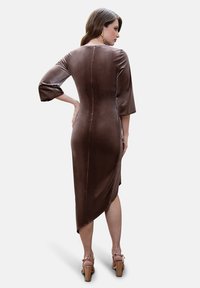 Brown velvet dress with elbow-length sleeves, fitted silhouette, and an asymmetrical hem; worn with wooden-heeled sandals.