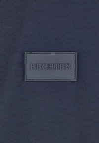 Marinefarbener Textiloberfläche mit einem strukturierten, schwarzen rechteckigen Patch, der das geprägte Wort "HECHTER" in fetter Schrift zeigt.