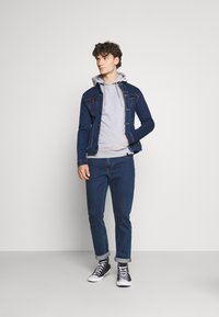 Denim jakna v temno modri barvi s svetlimi šivi, čez sivo kapuco. Slim-fit kavbojke v temno modri barvi, v kombinaciji s črnimi visoko-top čevlji.