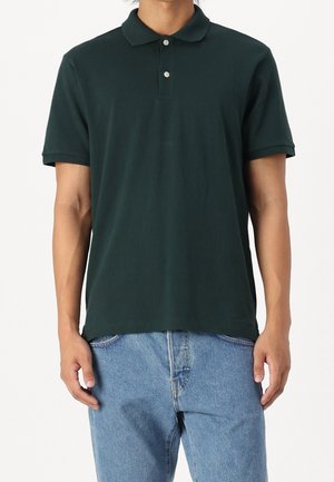 Mørkegrøn polo-shirt lavet af bomuld, med krave og to knapper, korte ærmer og lige bund. Bæret med blå jeans.