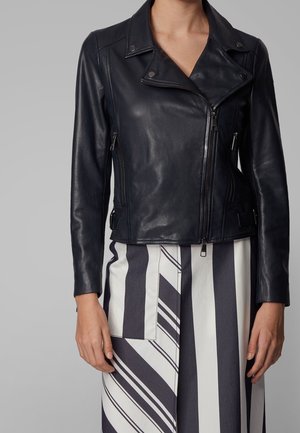 Personne portant une veste de motard en cuir noir avec une fermeture éclair asymétrique et une jupe à rayures verticales noires et blanches.