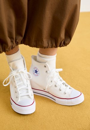 Osoba nosząca białe trampki Converse All Star za kostkę z białymi skarpetkami i brązowymi spodniami stojąca na żółtej podłodze.