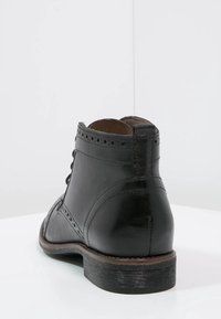 Bota de cuero negro hasta el tobillo con detalles perforados, textura suave, puntera redondeada y un tacón bajo y apilado. Diseño de cordones funcional.
