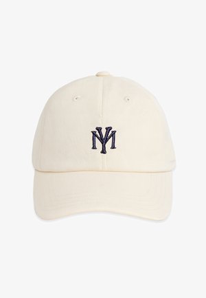 Jonge man met een beige pet met donker logo, lichtblauwe patroonige neksjaal, wit geribd shirt met logo en een donkere jas over de schouders gedrapeerd.