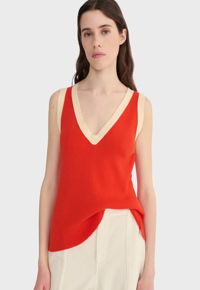 Femme portant un haut sans manches en tricot rouge avec une bordure beige et un pantalon blanc, se tenant devant un fond clair uni.