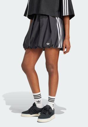 Personne portant une jupe noire Adidas avec des bandes blanches, des baskets noires et des chaussettes blanches Adidas, debout devant un fond clair uni.