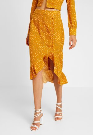 Jupe midi à pois jaune moutarde avec un ourlet volé et une fente sur le côté, comportant une rangée de boutons sur le côté. Associée à des talons blancs à lanières.