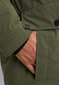 Olijfgroene jas met een klepzak, zichtbare stiksels en een logo-tag. Het materiaal lijkt lichtgewicht met een gladde textuur.