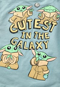 Hellblaues T-Shirt mit Cartoon-Charakteren, die grüne Haut und große Ohren haben, in braunen Roben gekleidet, mit dem Text "DIE SÜßESTEN DER GALAXIE."