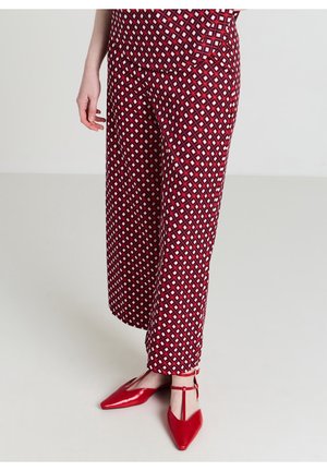 Femme portant un pantalon large à motif géométrique rouge et des ballerines rouges pointues avec bride en T, debout sur un sol clair devant un fond uni.