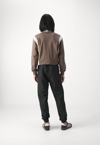 Brun beskuren sweatshirt med krämfärgade axelaccenter, parat med svarta avsmalnande byxor. Modellen visar en avslappnad bakvy. Sneakers med vita ränder.