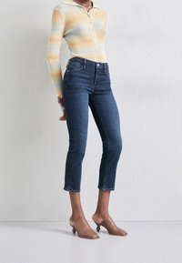 Suéter ligeramente rayado en amarillo y azul, con cuello y botones. Jeans azules de corte ajustado, de longitud recortada y con un ligero deshilachado en el dobladillo. Mules de tacón marrones.