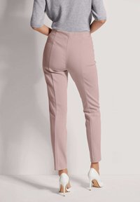 Helle rosa, enganliegende Hose mit glatter Textur, hoher Taille, vertikalen Nähten und weißen High Heels. Rückansicht vor einem neutralen Hintergrund.