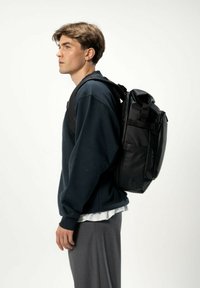 Svart ryggsäck med flera fack, justerbara remmar och en strömlinjeformad design. Bärs på ena axeln av en person i en marinblå sweatshirt.