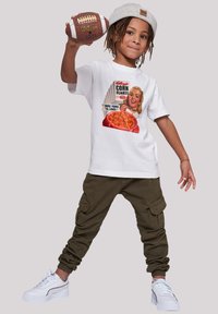 White T-shirt featuring a vintage Kellogg's Corn Flakes print, paired with olive cargo pants and white sneakers. Child holds a brown football.