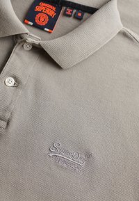 Jasnoszary polo wykonany z teksturowanej tkaniny, z dwoma guzikami i haftowanym logo "SuperDry" w pobliżu dolnego brzegu. Metki są w ciemnogranatowym i pomarańczowym kolorze.