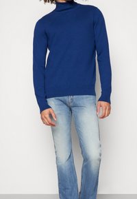 Suéter de cuello alto de canalé azul con mangas largas, combinado con jeans de denim descoloridos en azul claro. El tejido parece suave y ajustado.