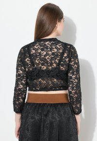 Schwarzes Spitzen-Crop-Top mit langen Ärmeln und floralem Muster. Der Rücken hat ein transparentes Design, kombiniert mit einem Rock mit braunem, elastischem Bund.