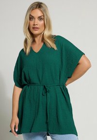 Green textured tunic with a V-neckline, short sleeves, and a tied waist. Features a relaxed fit and length reaching the hips.