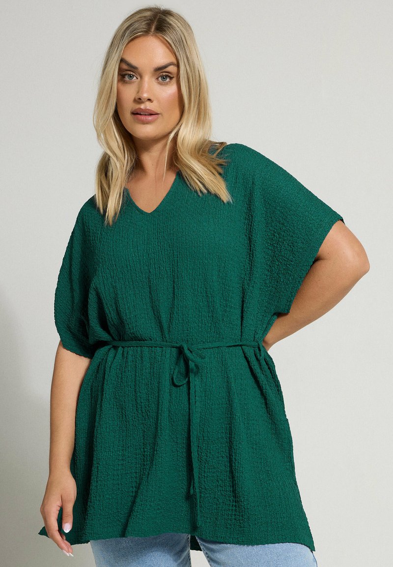 Green textured tunic with a V-neckline, short sleeves, and a tied waist. Features a relaxed fit and length reaching the hips.