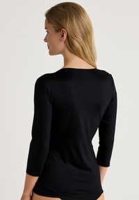 Zwarte ¾ mouw top met een gladde textuur, ronde halslijn en een getailleerde pasvorm. Van opzij weergegeven, met zicht op de achterkant.