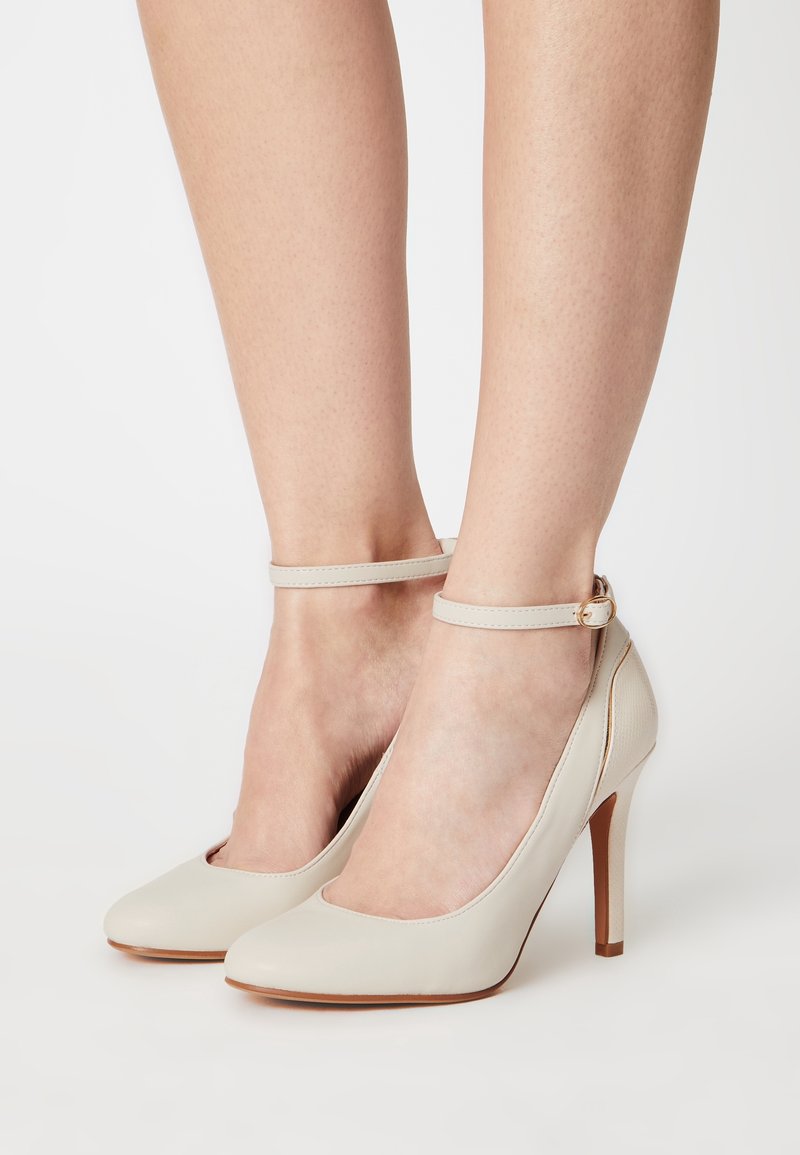 Chaussures à talons hauts beige en cuir lisse, à bout arrondi, avec bride autour de la cheville et talon aiguille fin, sur un fond neutre.