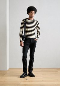 Svart og beige stripete langermet skjorte, svarte jeans, svart veske, og blanke svarte sko på en person som står på lyse tremgulv.