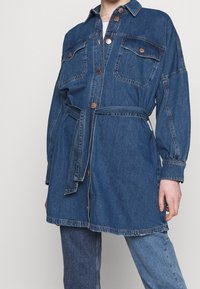 Jeansskjolklänning i mörkblått, med en knapplåsning framtill, två bröstfickor, långa ärmar och ett knytskärp i midjan. Kopparknappar accentuerar designen.