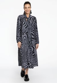 Langes schwarz-weißes Faltenkleid mit einem wellenförmigen Muster, Knopfverschluss und gebundenem Kragen, kombiniert mit schwarzen Plateauschuhen.