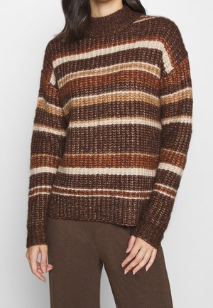 Femme portant un pull en maille rayé marron, orange et crème avec col, poignets et ourlet côtelés, associé à un pantalon côtelé marron.