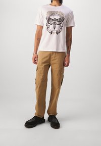 Camiseta blanca con estampado gráfico y logo, combinada con pantalones cargo beige con bolsillos laterales. El conjunto se completa con zapatos negros.