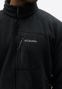 Chaqueta de forro polar negra Columbia con cremallera frontal y bolsillo en el pecho con cremallera, llevada por una persona con la parte inferior del rostro y el cuello visibles.
